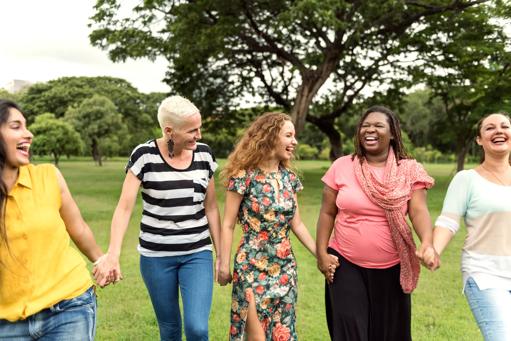 Diverse group of women laughing and holding hands outdoors, symbolizing community and wellness